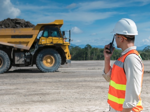 Man wearing a hard hat talking into a radio to someone driving a truck full of mined materials